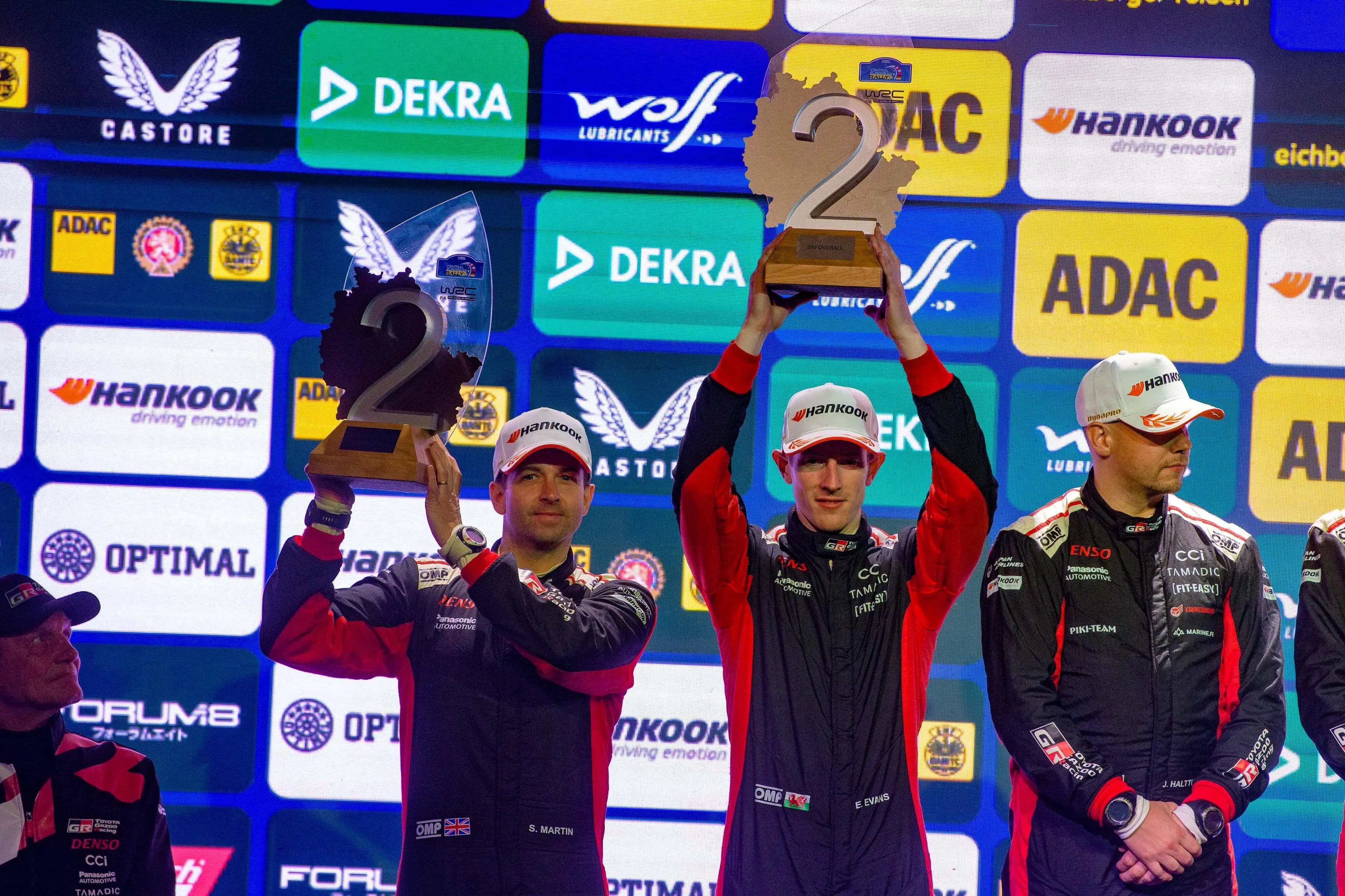 Elfyn Evans (centre) lifts his trophy for runners-up spot. Pic. Alamy